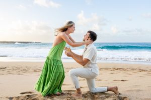 Man proposes to woman in green dress on sandy beach with ocean waves in background.