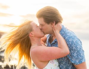 Couple sharing passionate kiss at sunset on beach with golden light and wind-blown hair.