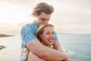 Young couple embracing at sunset by the ocean, woman smiling joyfully in golden light.