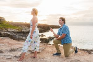 Man proposing on one knee to surprised woman on rocky coastal cliff at sunset in Hawaii.