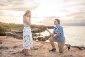 Man on one knee proposing to emotional woman on rocky oceanside cliff at golden sunset.