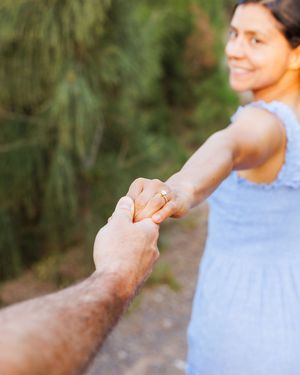 Close-up of woman showing engagement ring while holding mans hand outdoors with greenery.