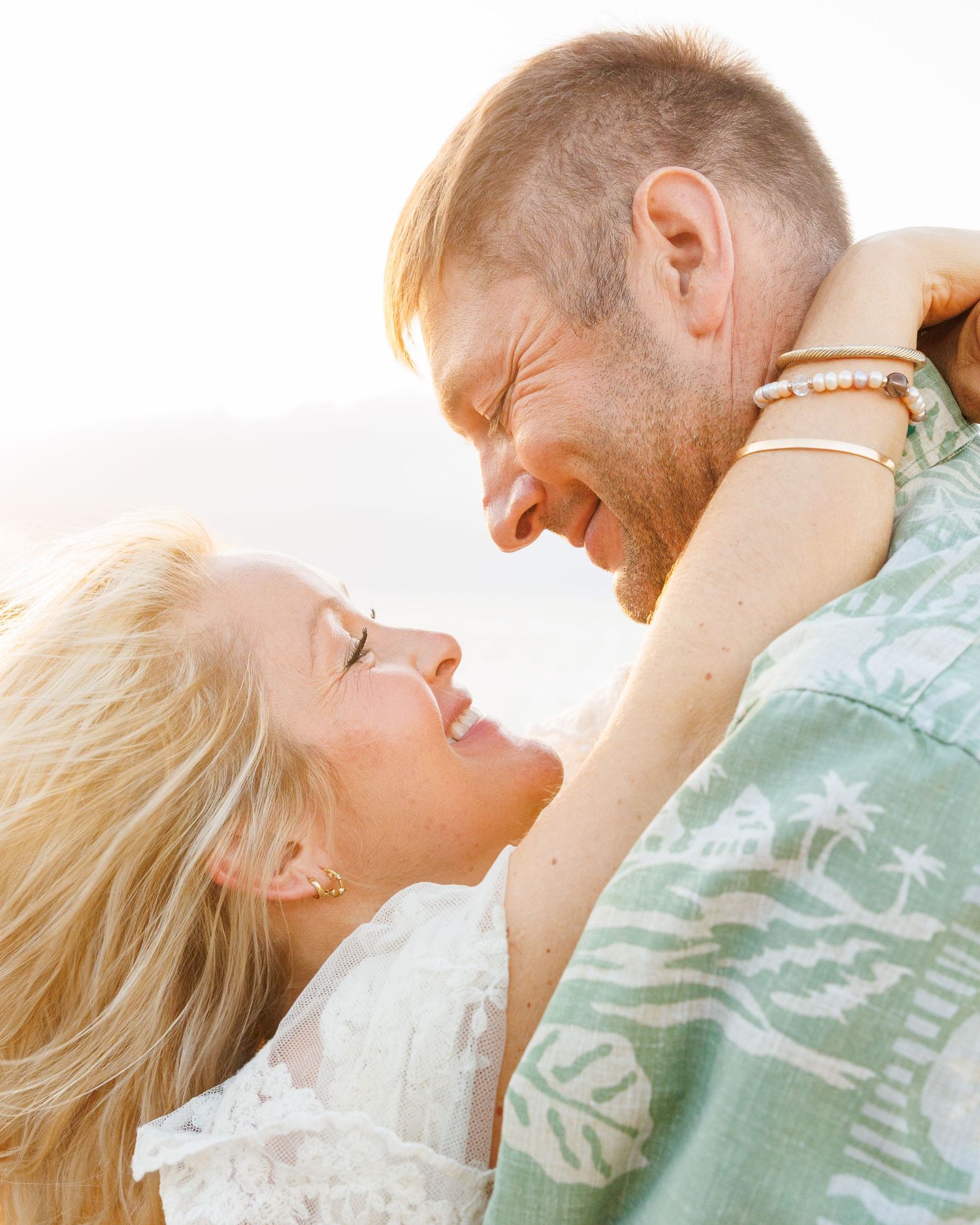 Romantic couple gazes lovingly at each other in soft golden light on a sun-drenched beach.