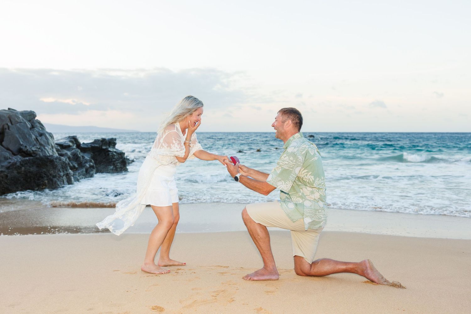 Man proposing on one knee to woman on sandy beach at sunset with ocean waves in background.