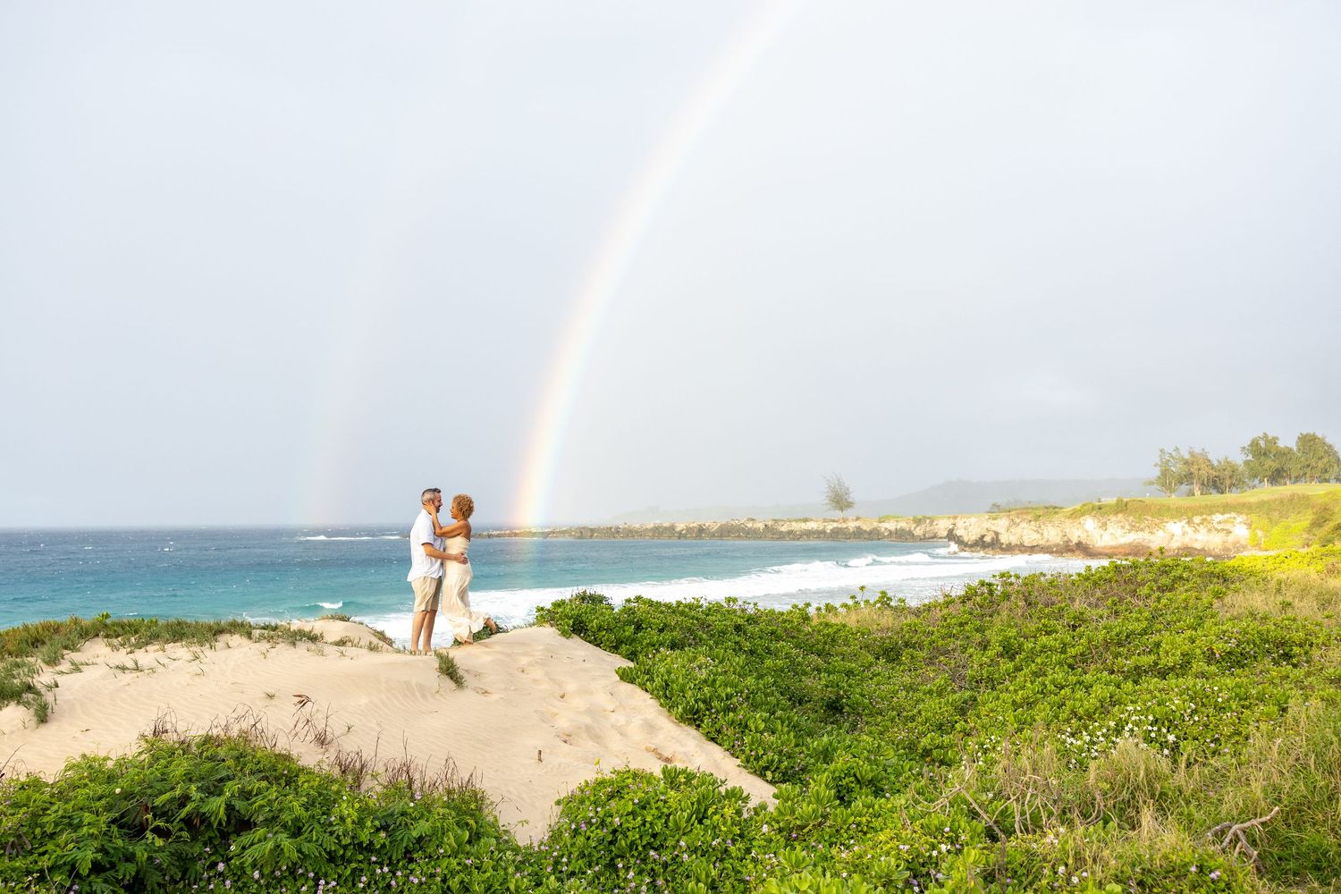 Couple standing on sand dunes with a stunning double rainbow over the ocean.