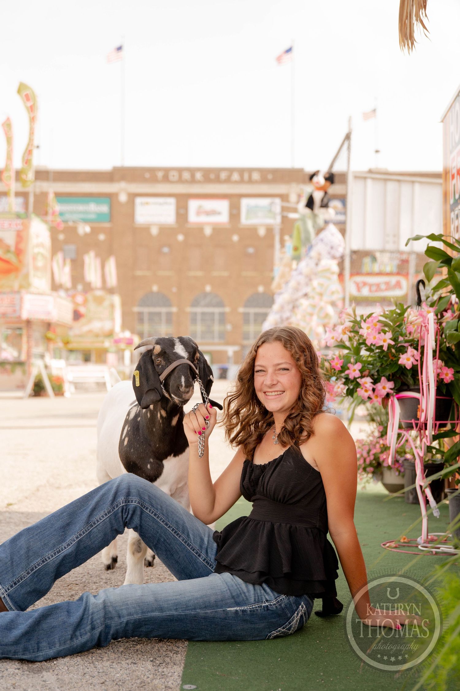 Smiling teenage girl sitting with a black and white goat at the York Fair, surrounded by pink flowers and fair buildings.
