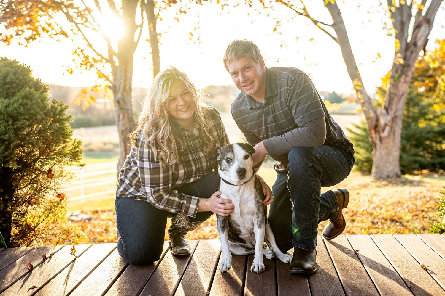 Smiling couple crouching with their dog on a wooden deck in fall.