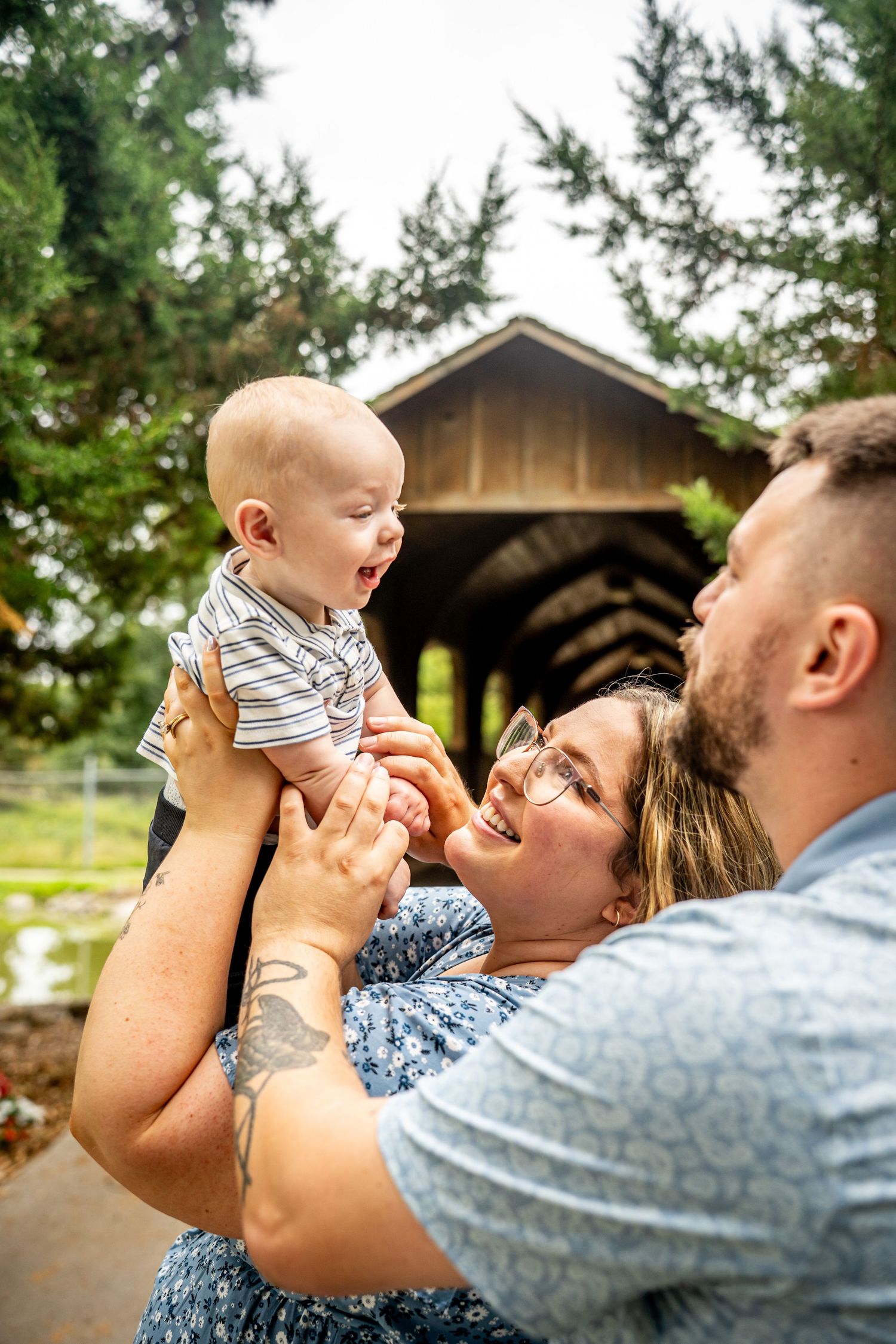 A happy couple holds their laughing baby outdoors near a rustic covered bridge surrounded by lush green trees.