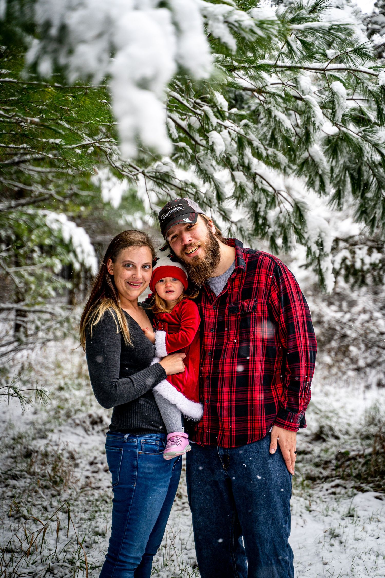 Young family of three posing outdoors among snow-covered pine trees in winter.