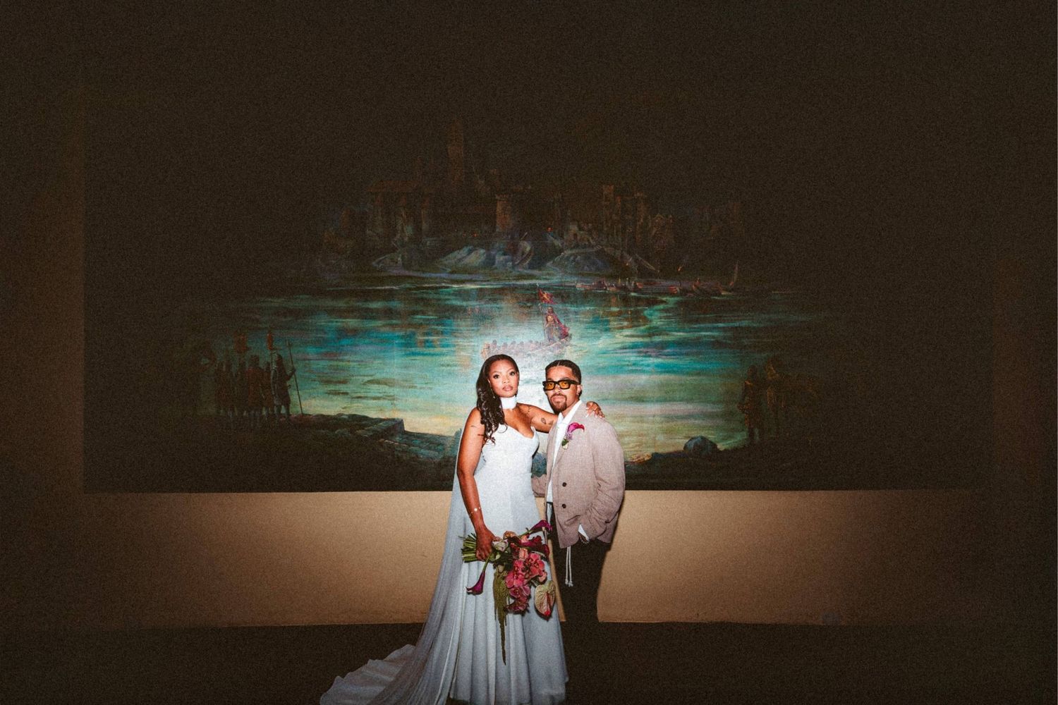 Bride and groom posing elegantly in front of a large colorful painting at their wedding reception.