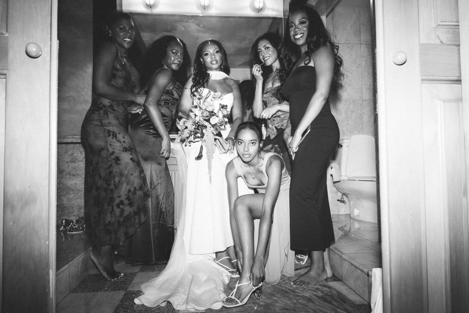 Black and white photo of a bride holding a bouquet surrounded by five bridesmaids posing together in a doorway.
