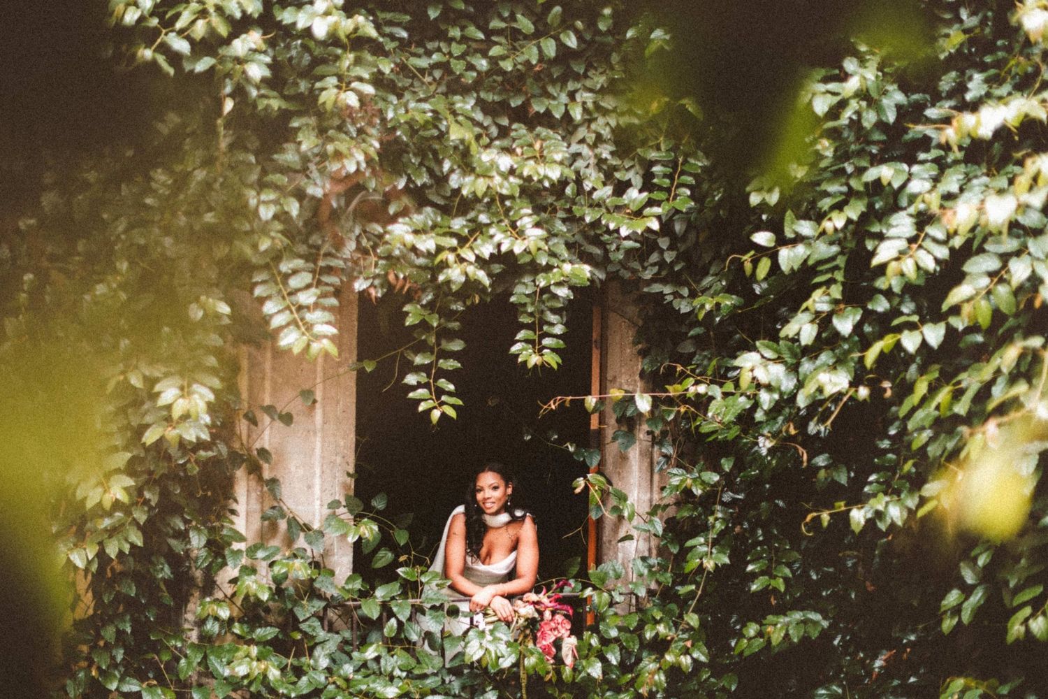 Bride surrounded by lush green ivy framing a rustic window in a romantic outdoor wedding portrait.