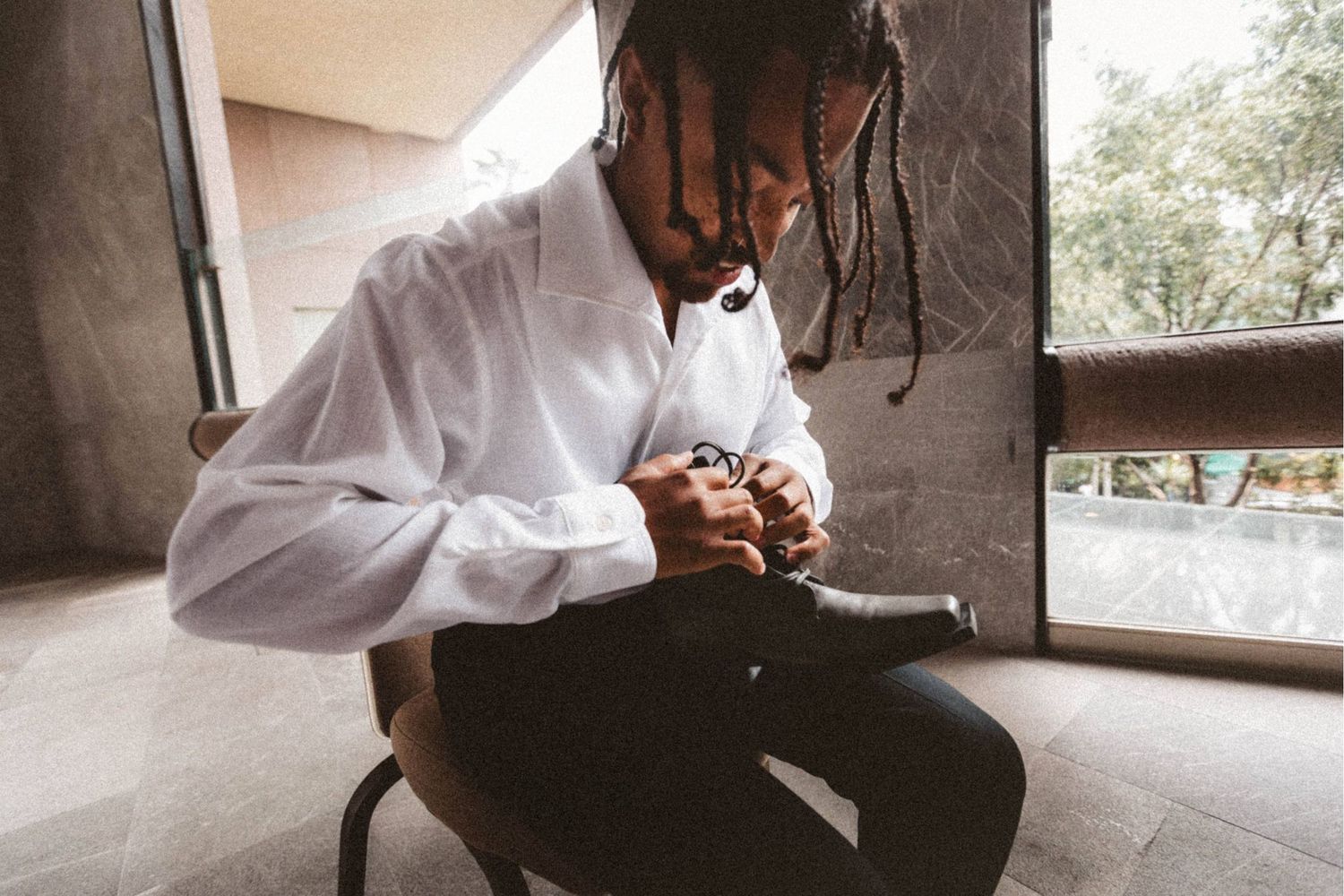 A man with dreadlocks wearing a white shirt sits on a chair, focused on adjusting something in his hands.