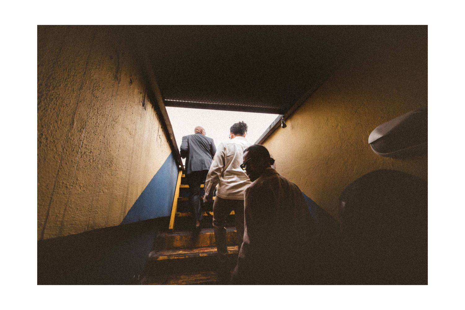 People ascending dark stairwell with yellow railing in urban setting.