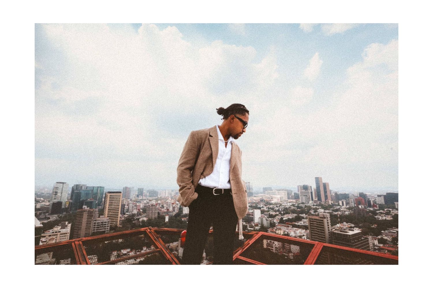 Man in blazer stands on rooftop edge with city skyline and cloudy blue sky behind him.