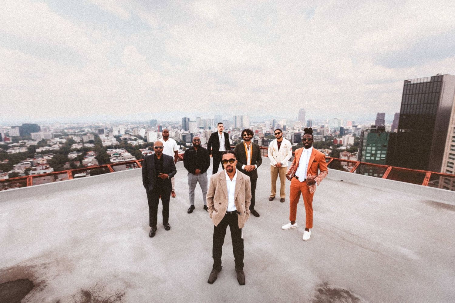 Group of stylishly dressed men posing on a rooftop with a city skyline backdrop under cloudy skies.