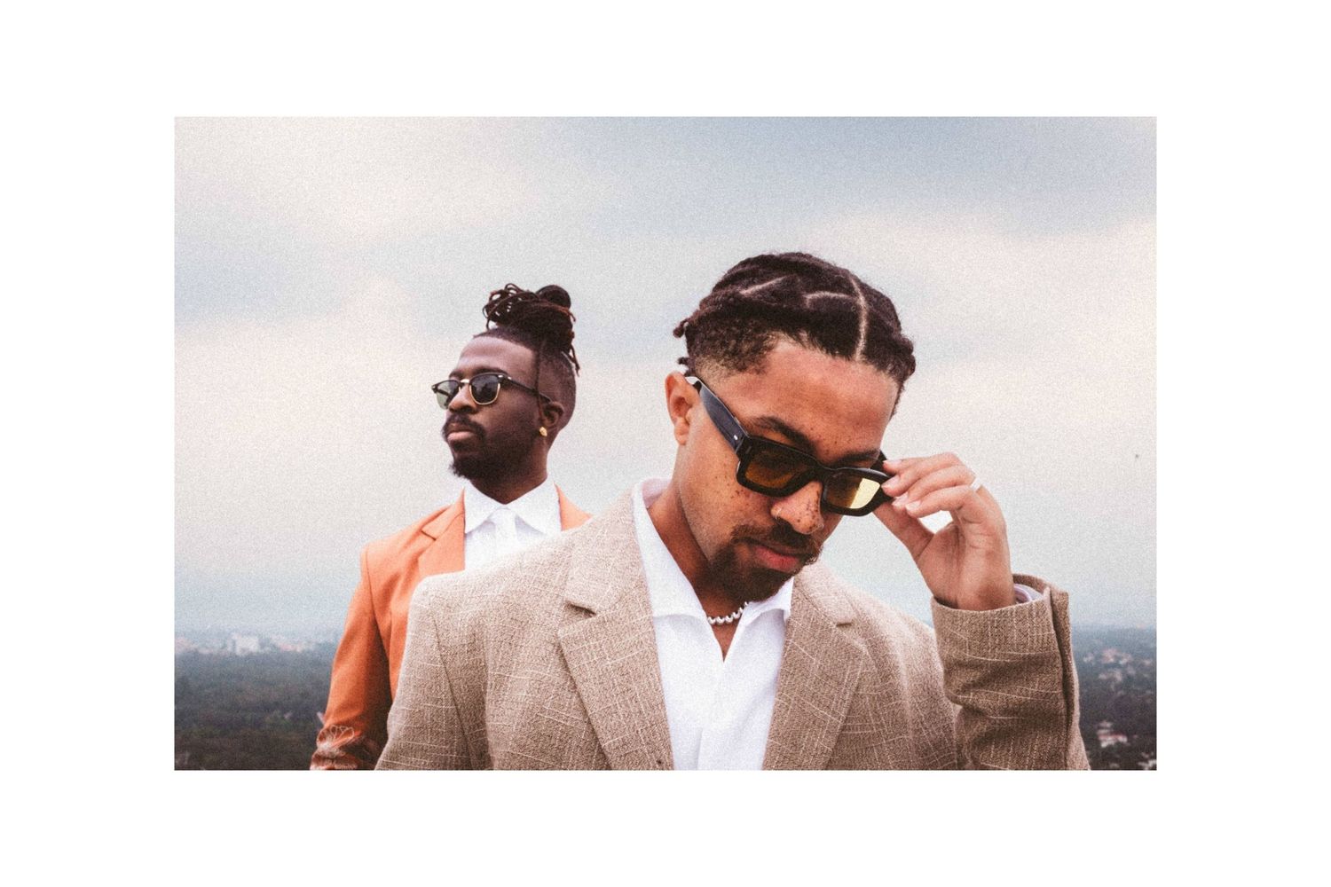 Two stylish men in tailored suits pose outdoors, one wearing sunglasses and braids, against a city skyline backdrop.