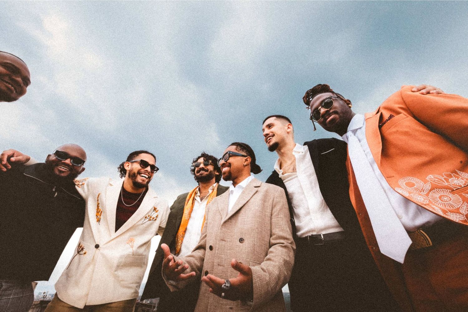 Six stylishly dressed men posing together outdoors under a cloudy sky, wearing elegant suits in neutral and bold tones.