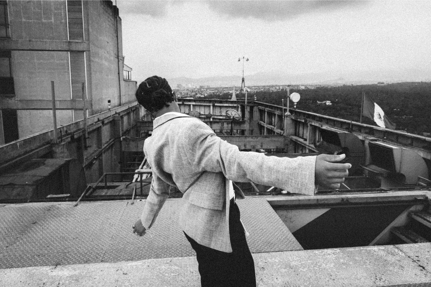 Black and white photo of a person standing on a rooftop overlooking an industrial cityscape, arms outstretched.