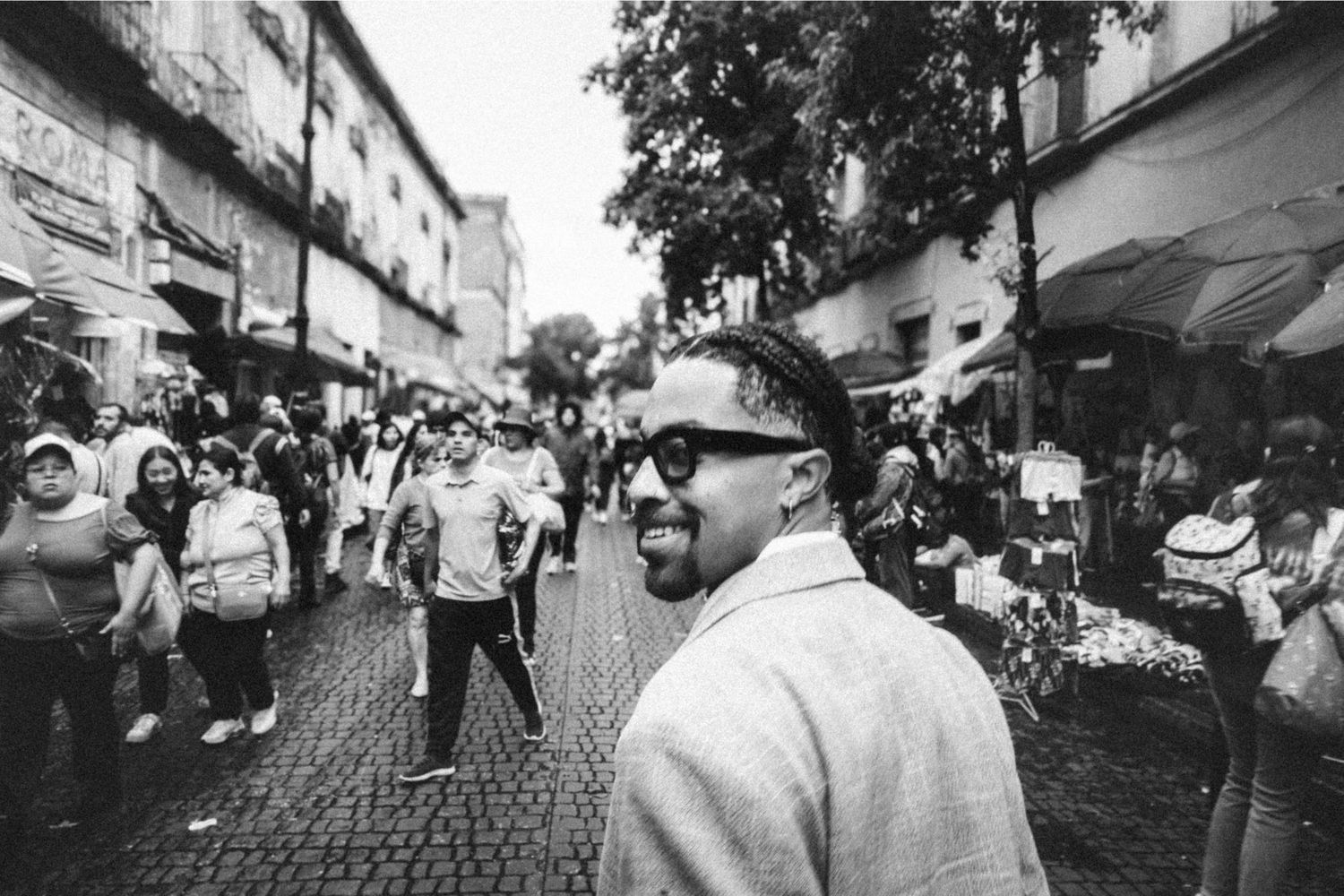 Man with sunglasses smiling on a busy cobblestone street market in black and white photo.
