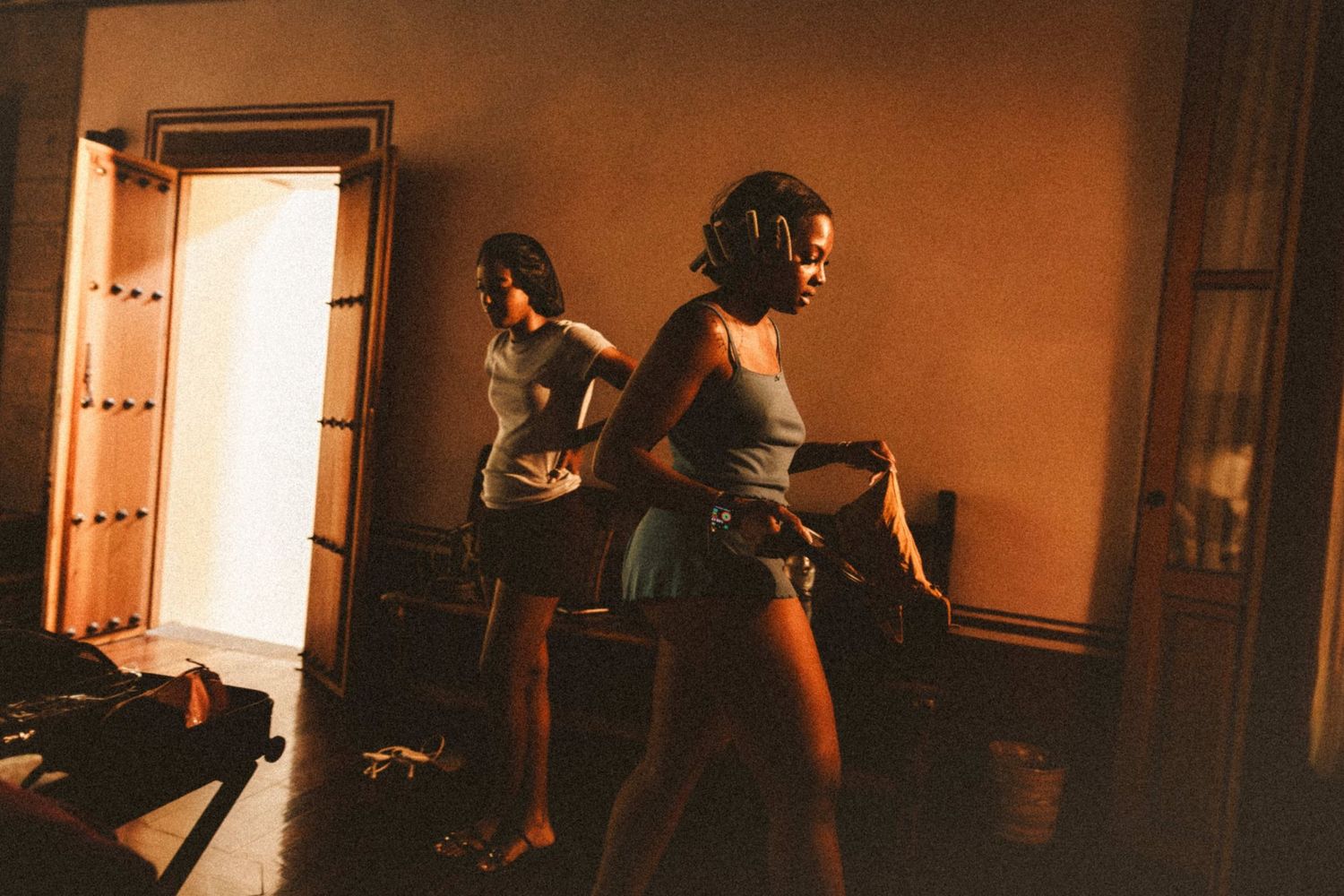 Two women in casual clothing stand in a warmly lit room near an open door in a candid indoor scene.