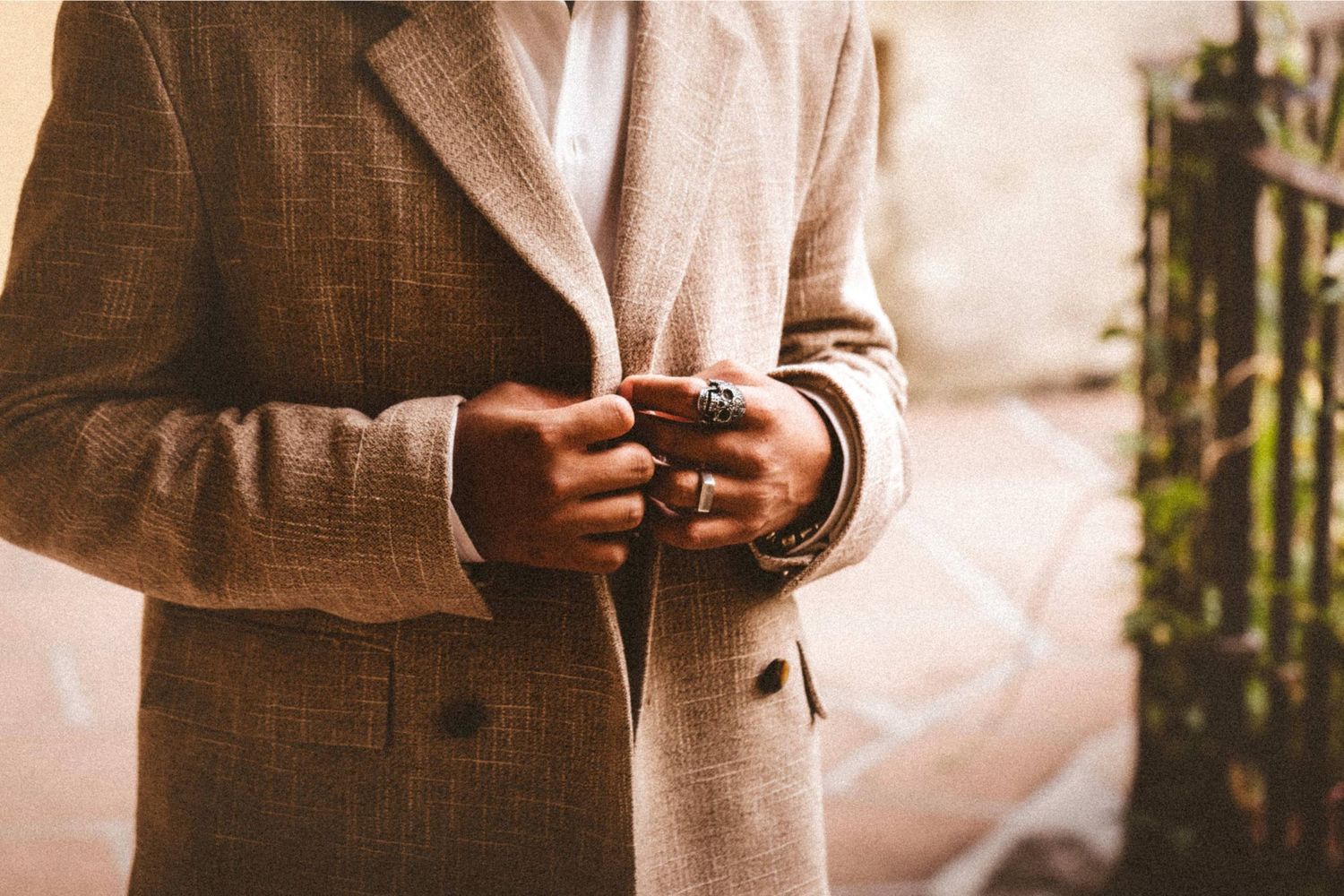 Man in a tailored beige plaid blazer holding hands together, wearing rings and bracelets, outdoors.