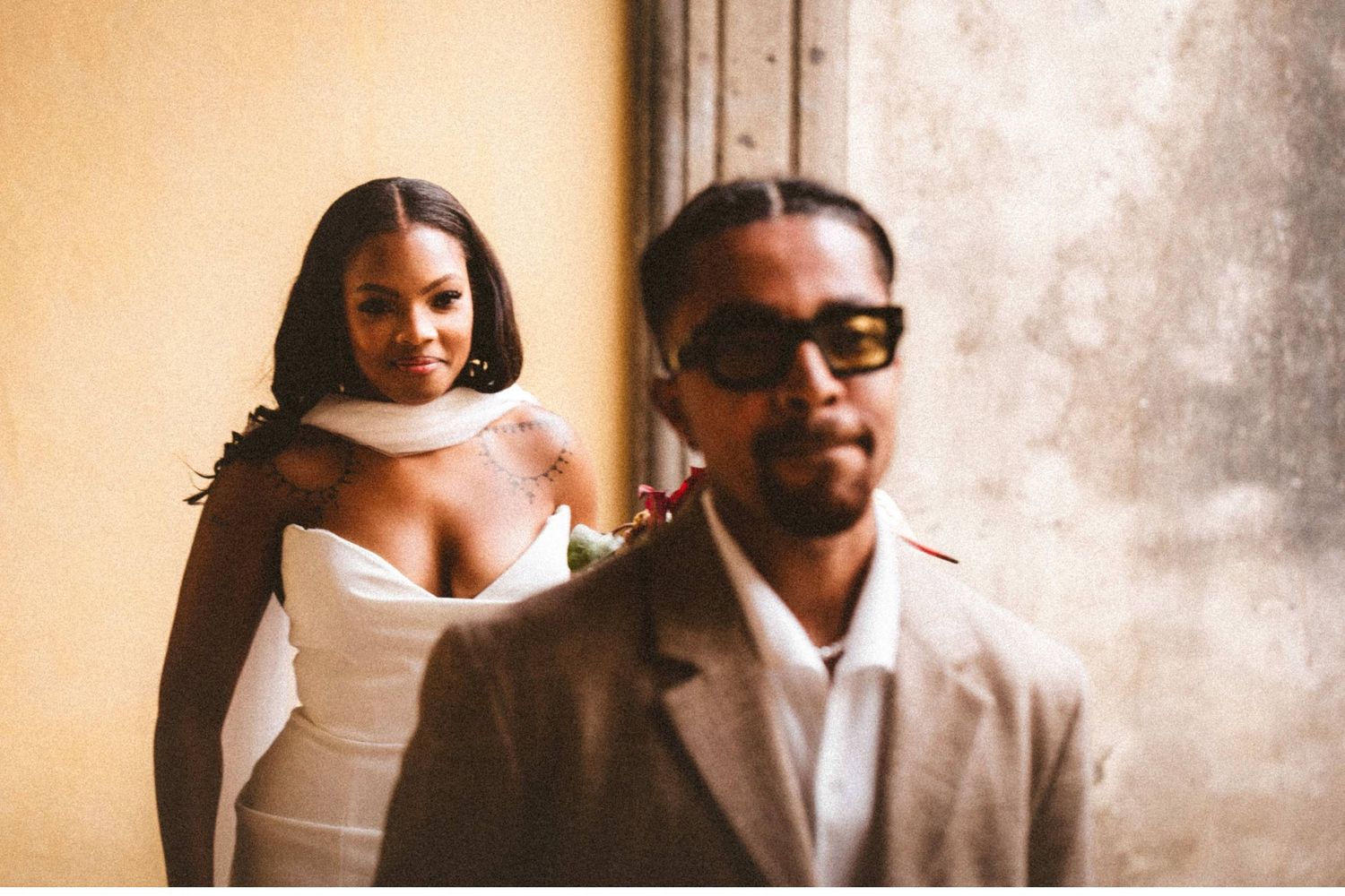 Elegant bride in white dress poses with groom in tan suit and sunglasses against a yellow wall.