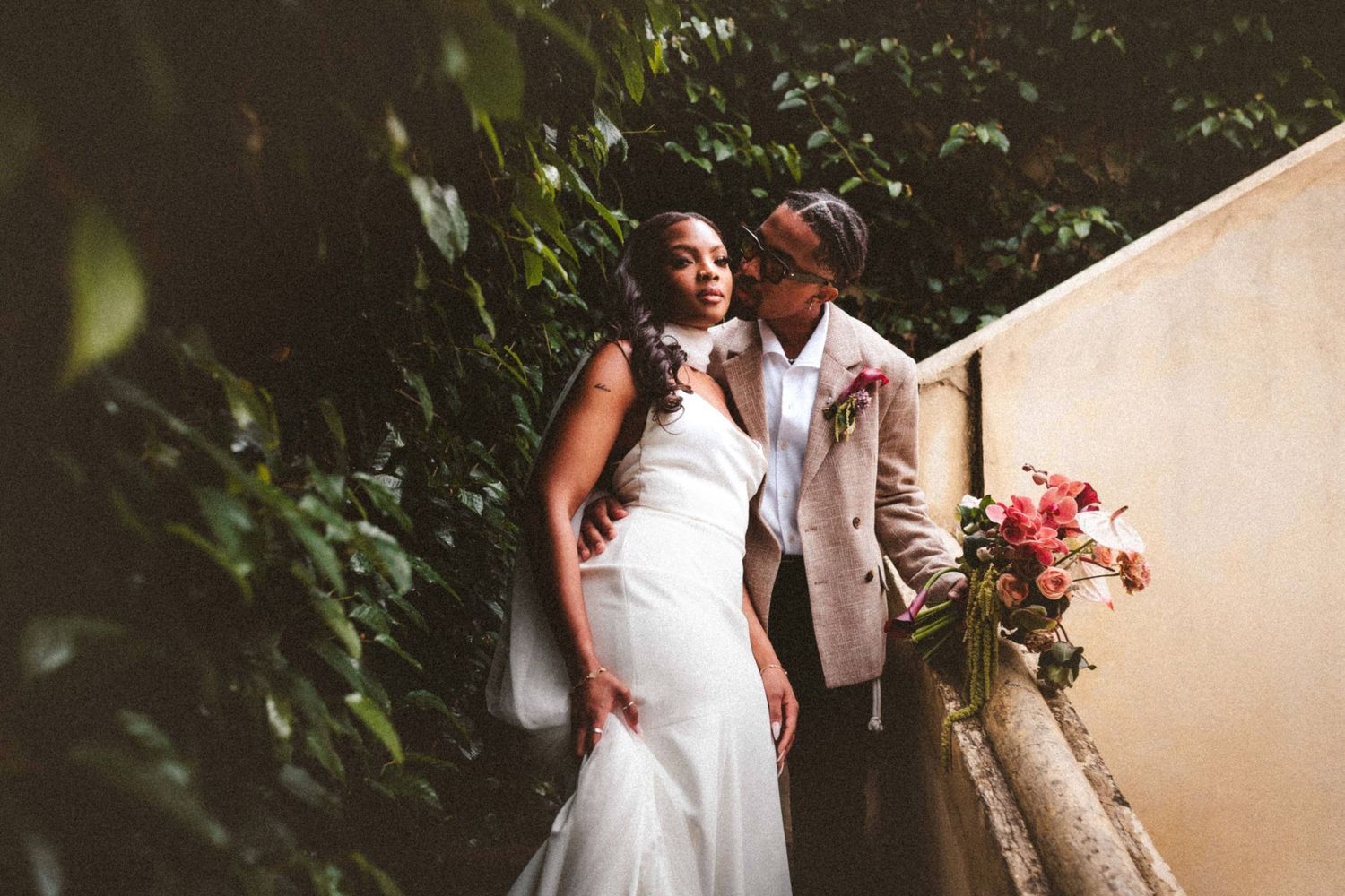 Black bride and groom pose elegantly beside lush green wall with pink bouquet.