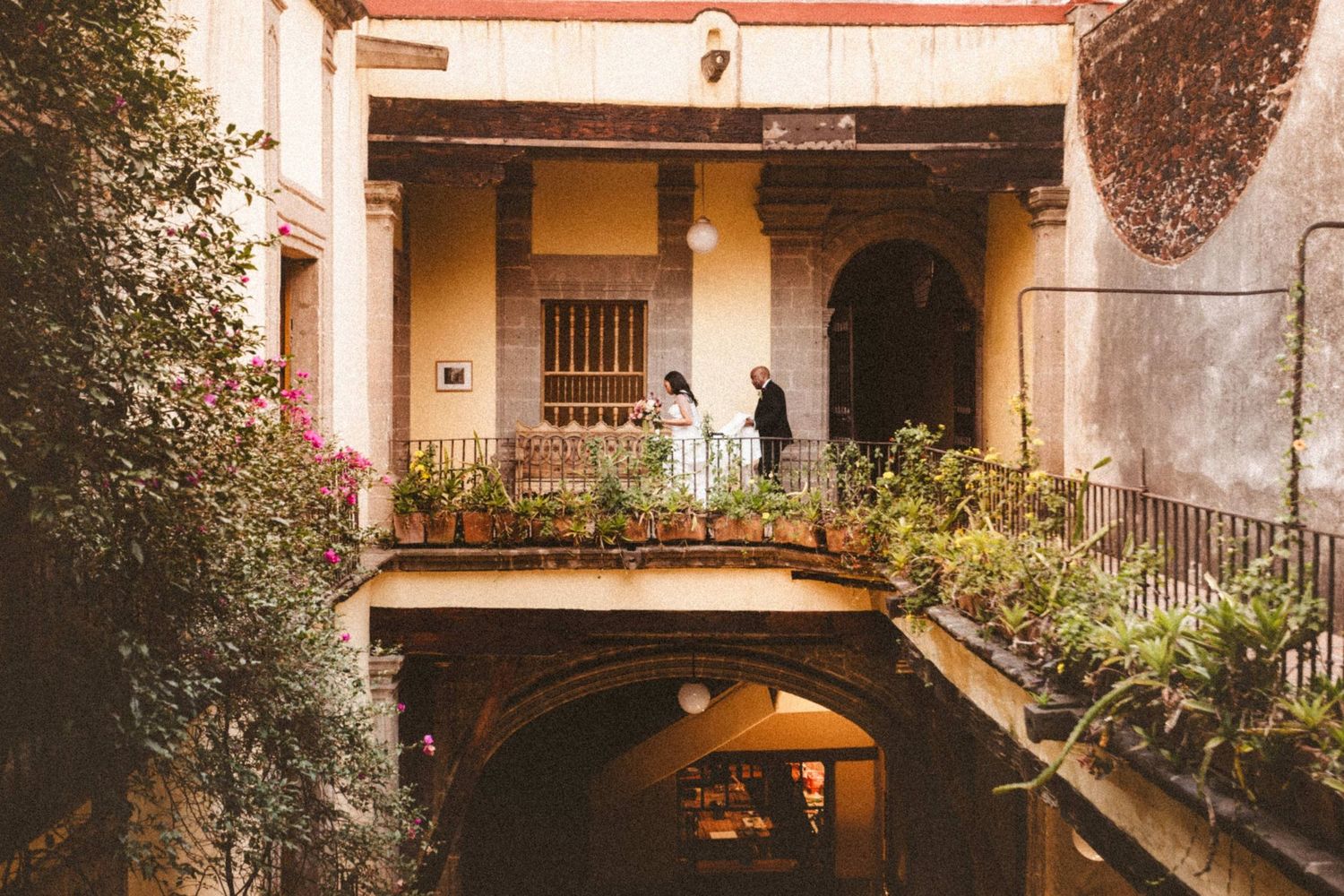 Couple on ornate balcony of historic courtyard venue with lush greenery, arched doorways, and warm Mediterranean architecture.