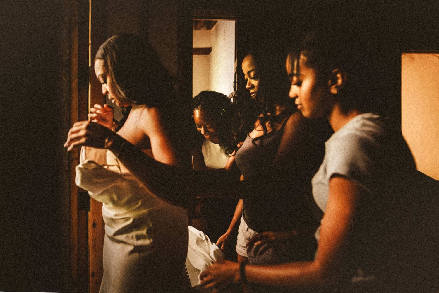 Bridesmaids helping a bride get dressed in a warmly lit room before the wedding ceremony.