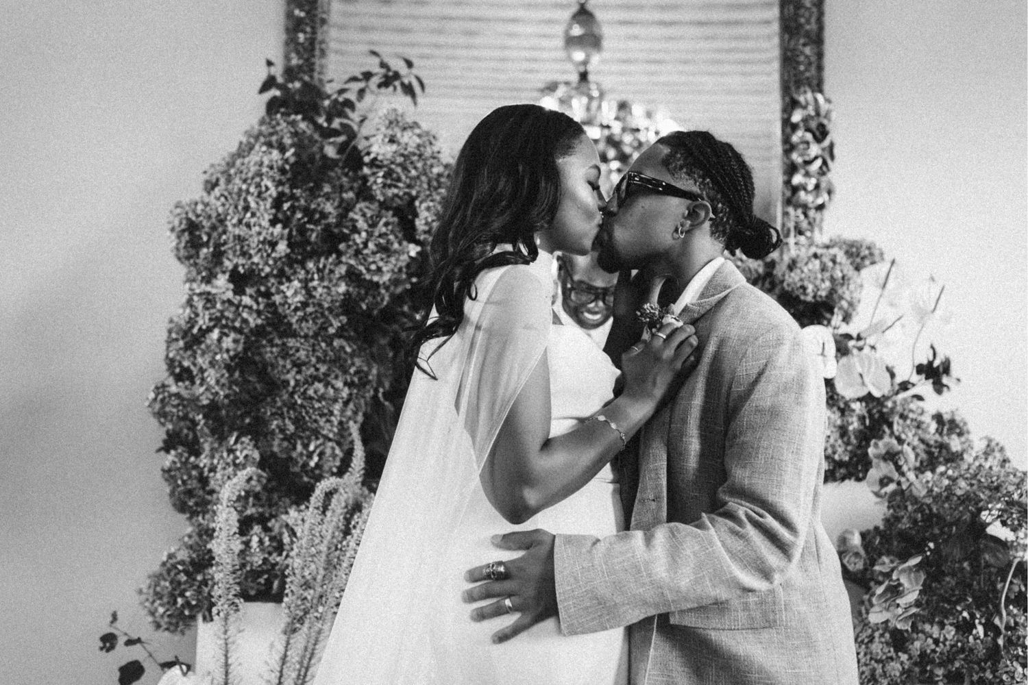 Black and white photo of a couple sharing a romantic kiss at their wedding ceremony surrounded by lush floral arrangements.