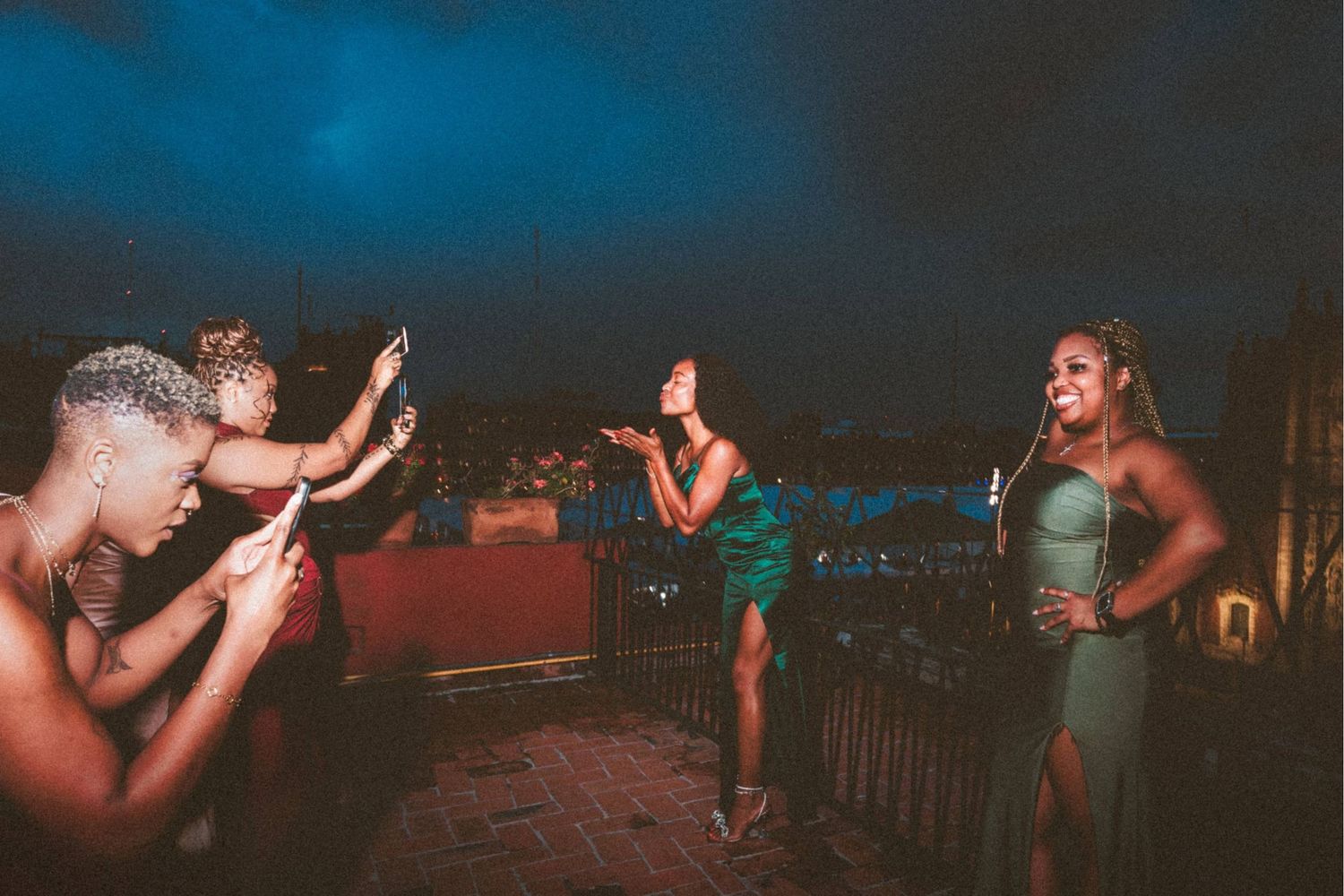 Four women celebrate on a rooftop at night, taking photos and dancing under a dramatic blue sky.