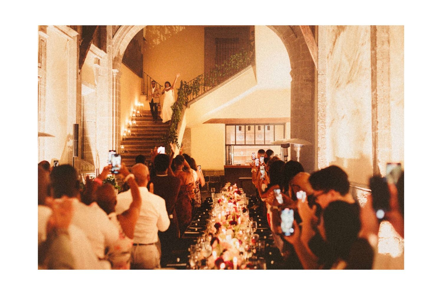 Elegant wedding ceremony in grand venue with floral aisle, guests holding phones, ornate staircase, and warm golden lighting.