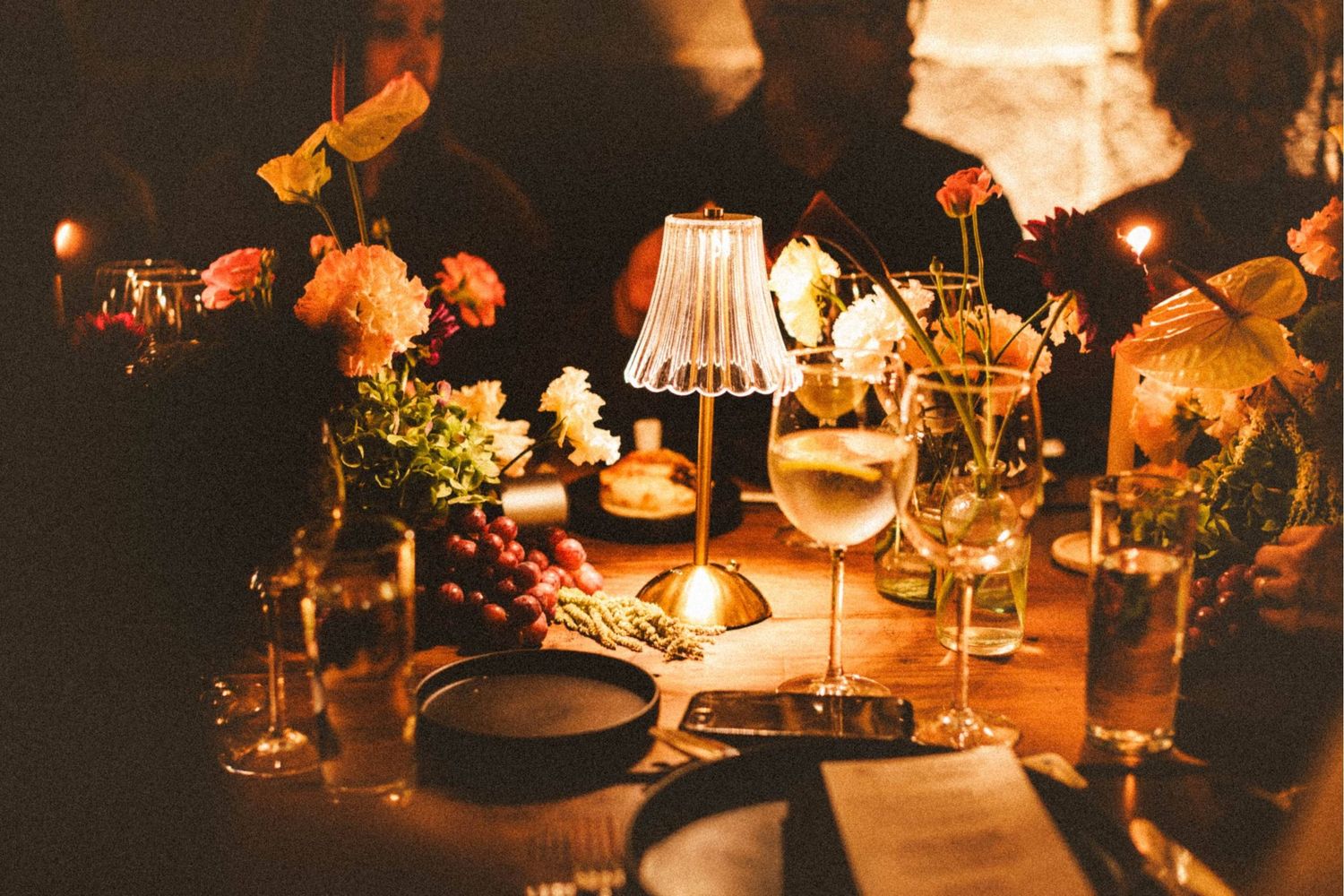 Romantic candlelit dinner table with floral arrangements, glowing lamp, and elegant glassware in warm amber light.