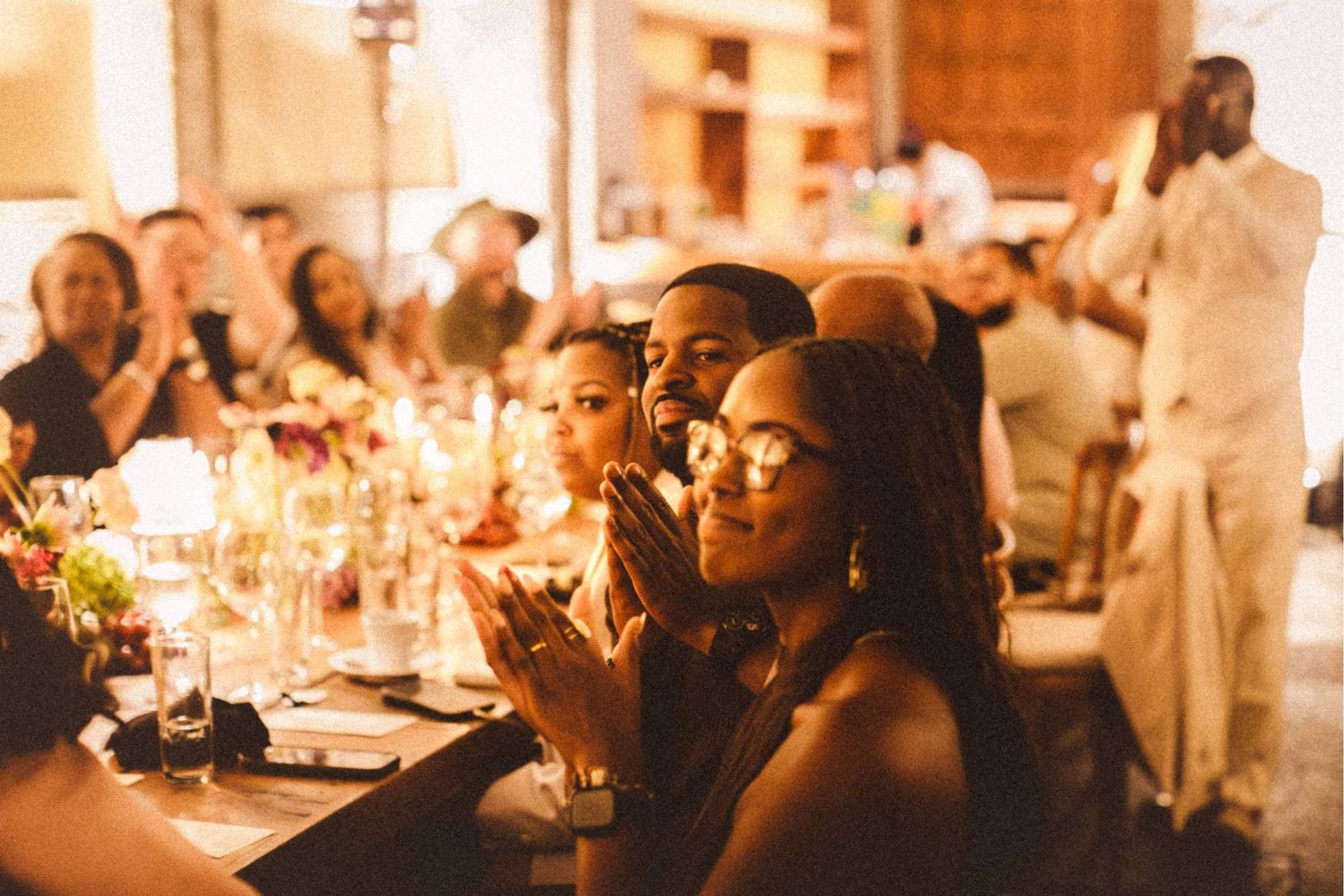 Guests clapping and celebrating at a warmly lit wedding reception dinner table with candles and flowers.