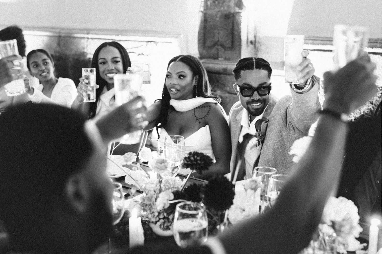 Black and white wedding reception photo of smiling couple toasting with guests at elegantly decorated table.