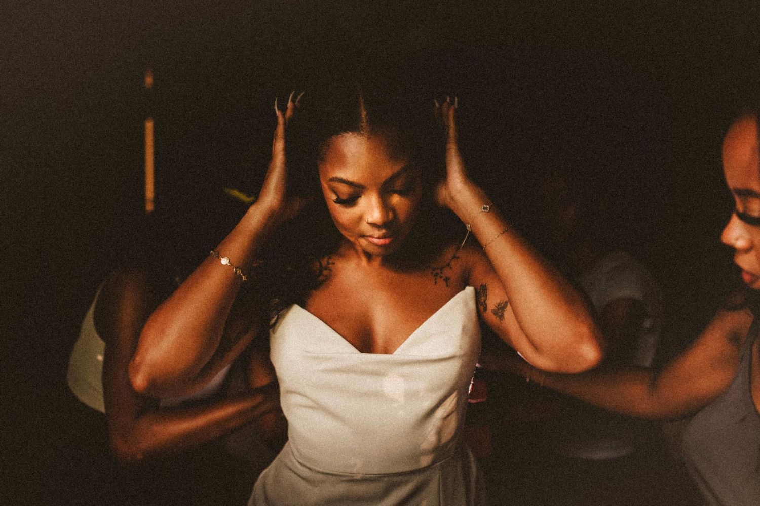 Bride in white strapless gown adjusts her hair with both hands, assisted by bridesmaids in dimly lit venue.