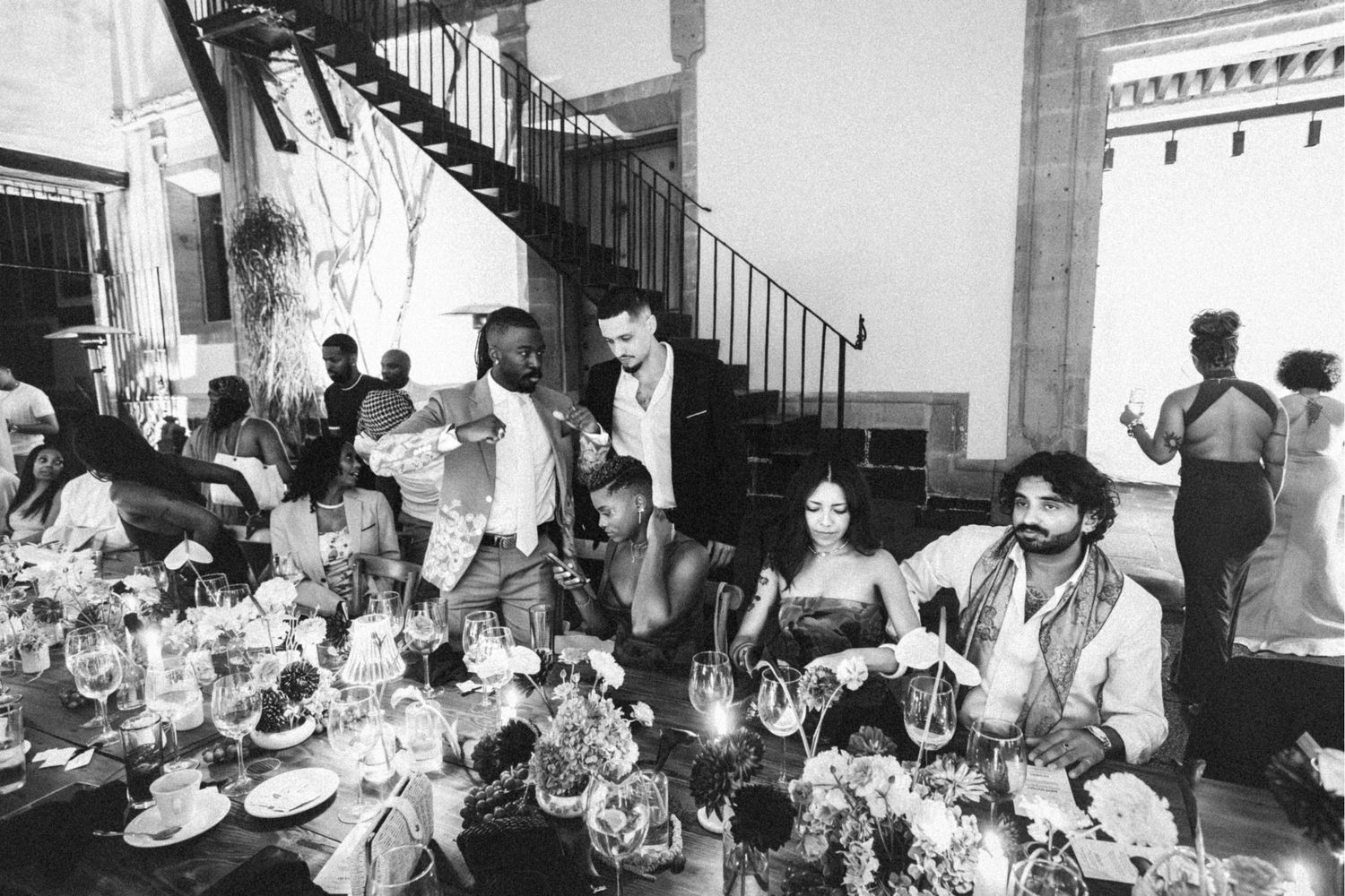 Black and white photo of elegantly dressed guests dining at a grand reception with spiral staircase.