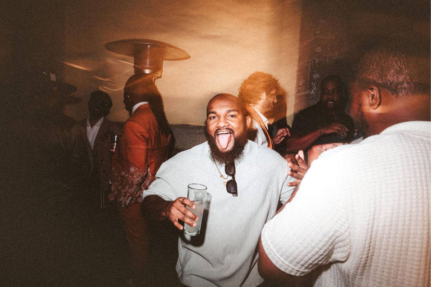 Man sticking tongue out holding drink at lively indoor party with guests in background.