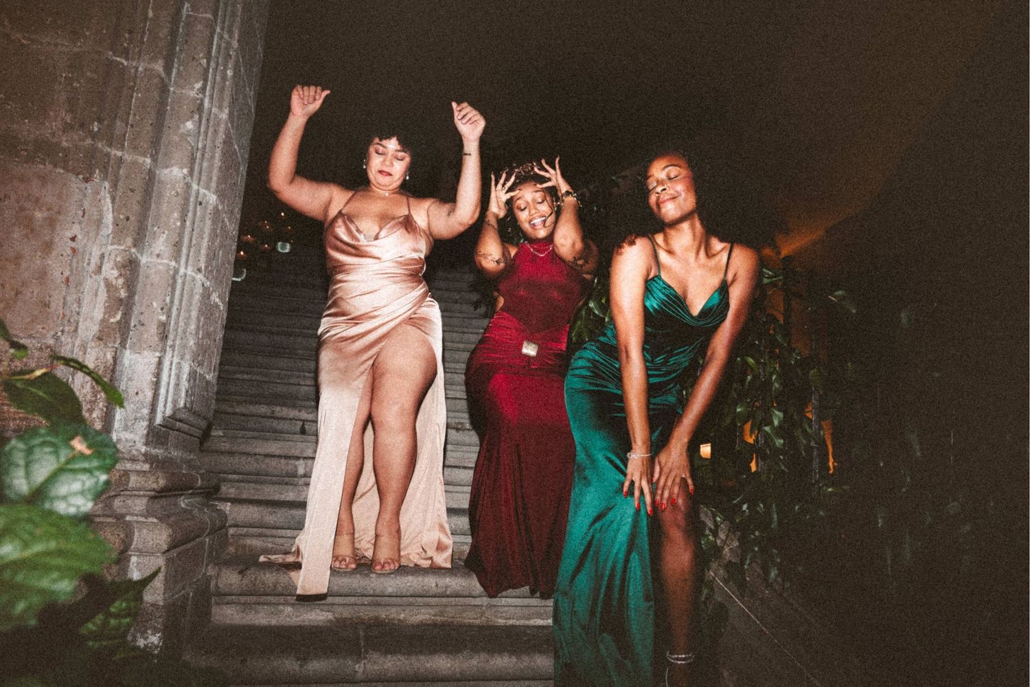 Three women in elegant gowns laughing and dancing on stone steps at a night event.