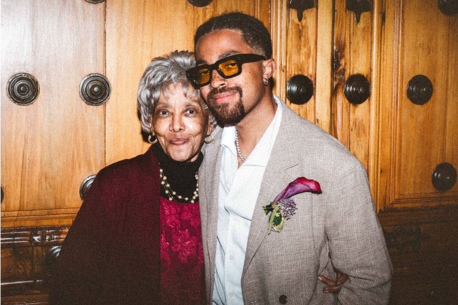 An elderly woman and a young man in a beige suit with orange sunglasses pose together indoors.