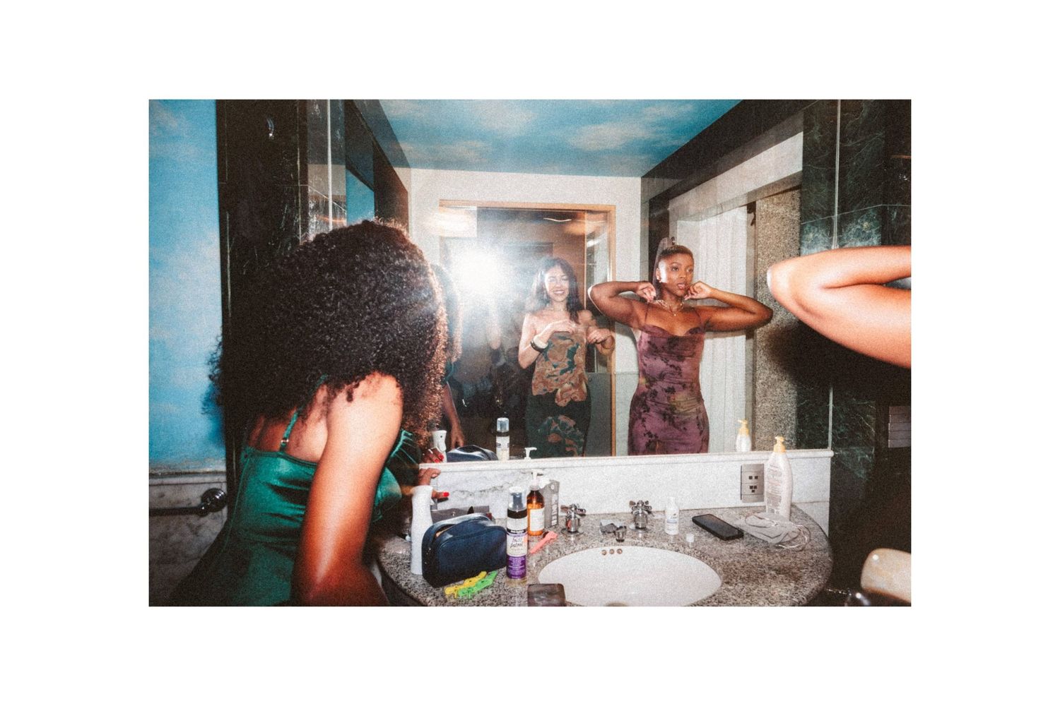 Women getting ready together in a bathroom mirror, capturing candid pre-wedding preparation moments.