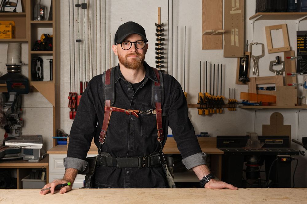 Bearded craftsman in black overalls with leather suspenders stands confidently in a well-equipped woodworking workshop.