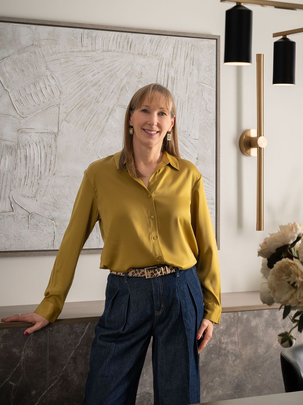 Woman in mustard yellow silk blouse and wide-leg jeans smiling in a modern, elegantly decorated interior.