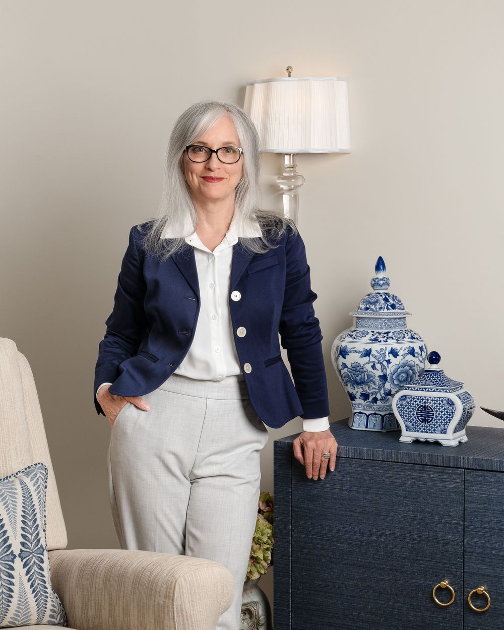 Elegant woman in navy blazer and gray trousers poses beside blue and white ginger jar on dark dresser.