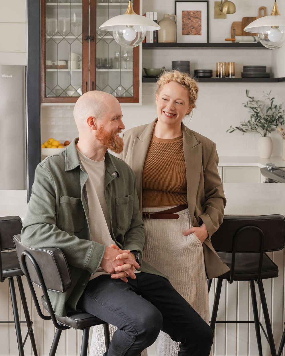 A smiling couple sits together in a modern kitchen, the man in a green jacket and woman in a tan blazer on black bar stools.