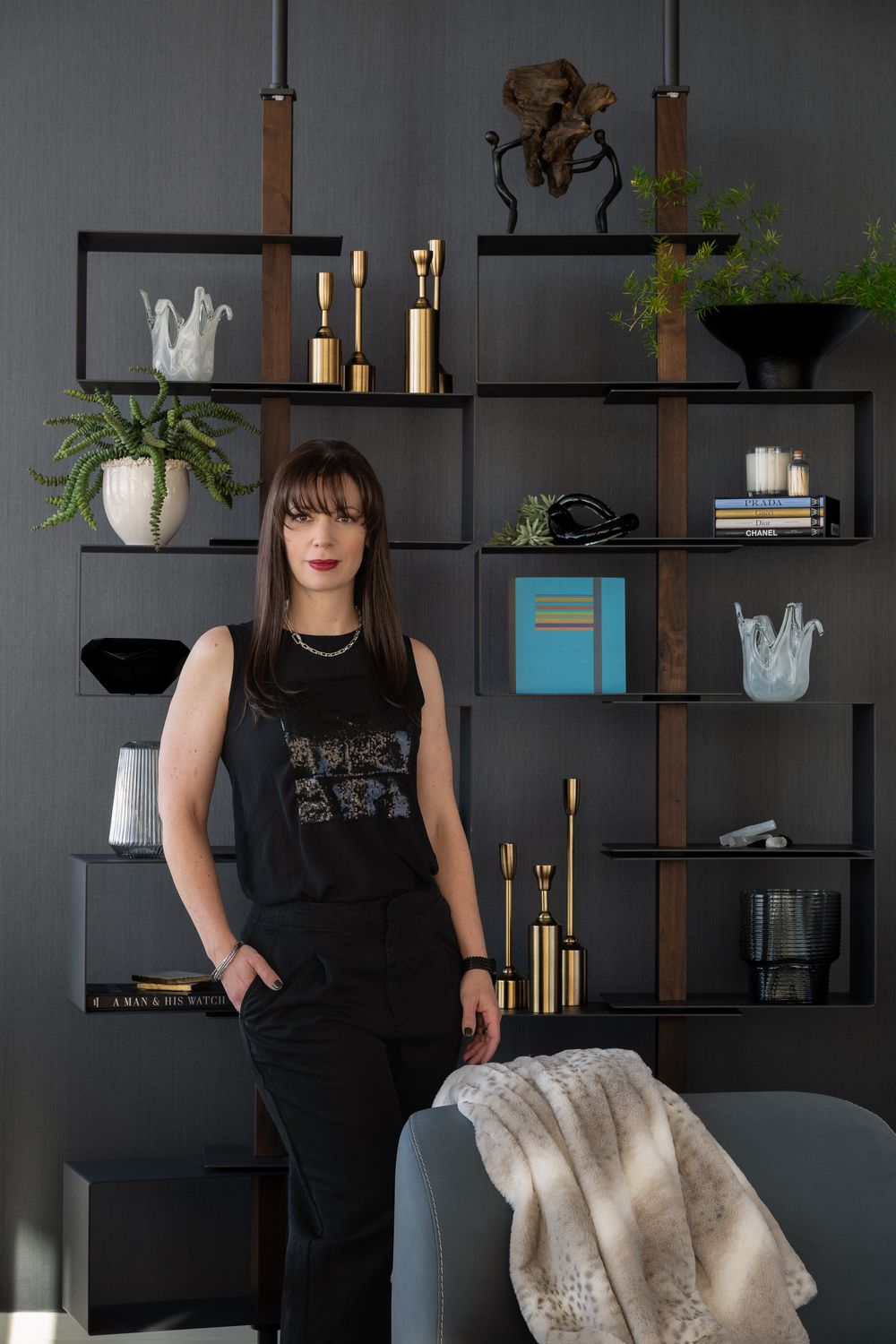 Woman in black outfit poses confidently in front of a stylish dark shelving unit with gold candles and decorative objects.