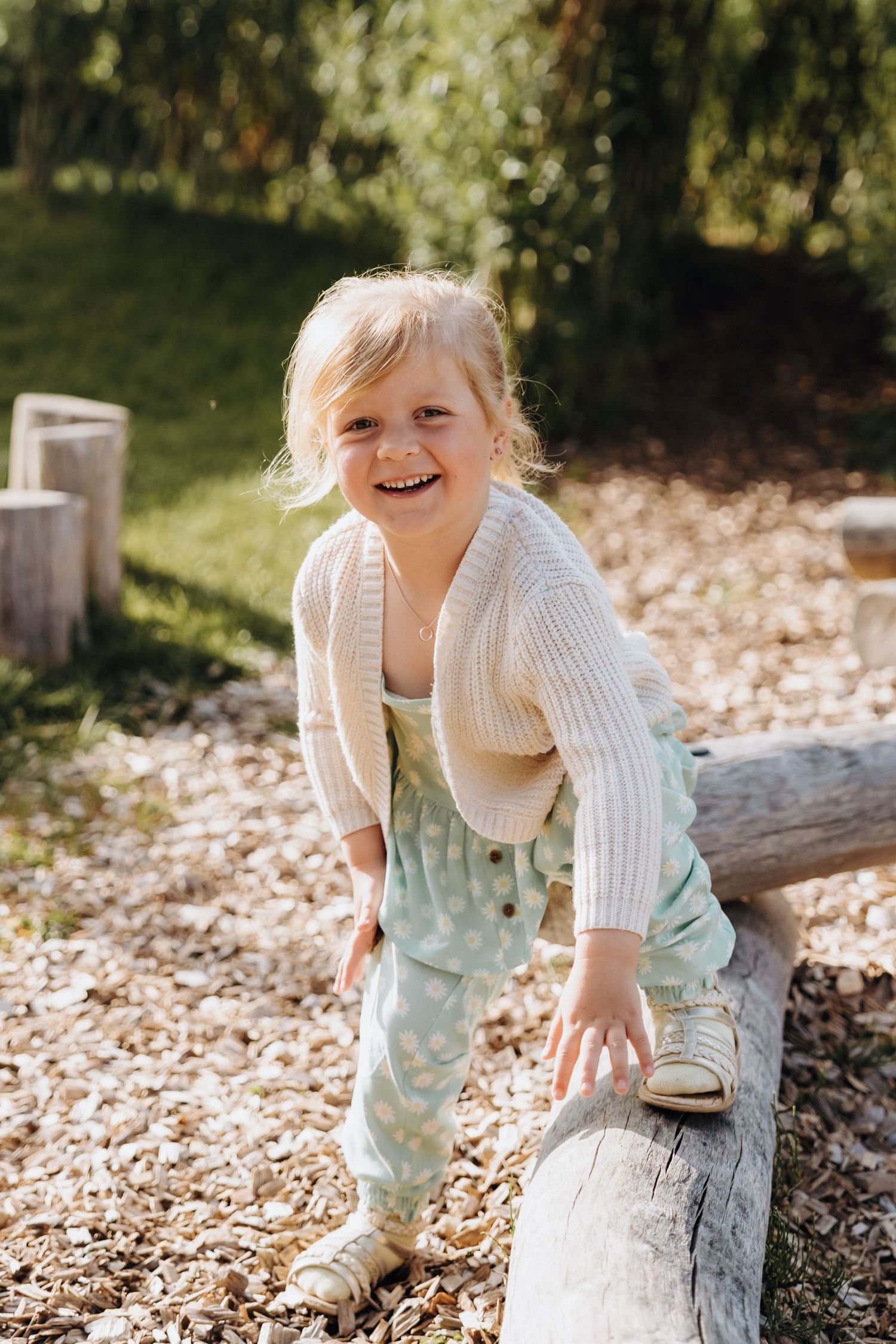 Happy toddler girl climbing on a log at an outdoor playground, wearing a cream knit cardigan and mint green pants.