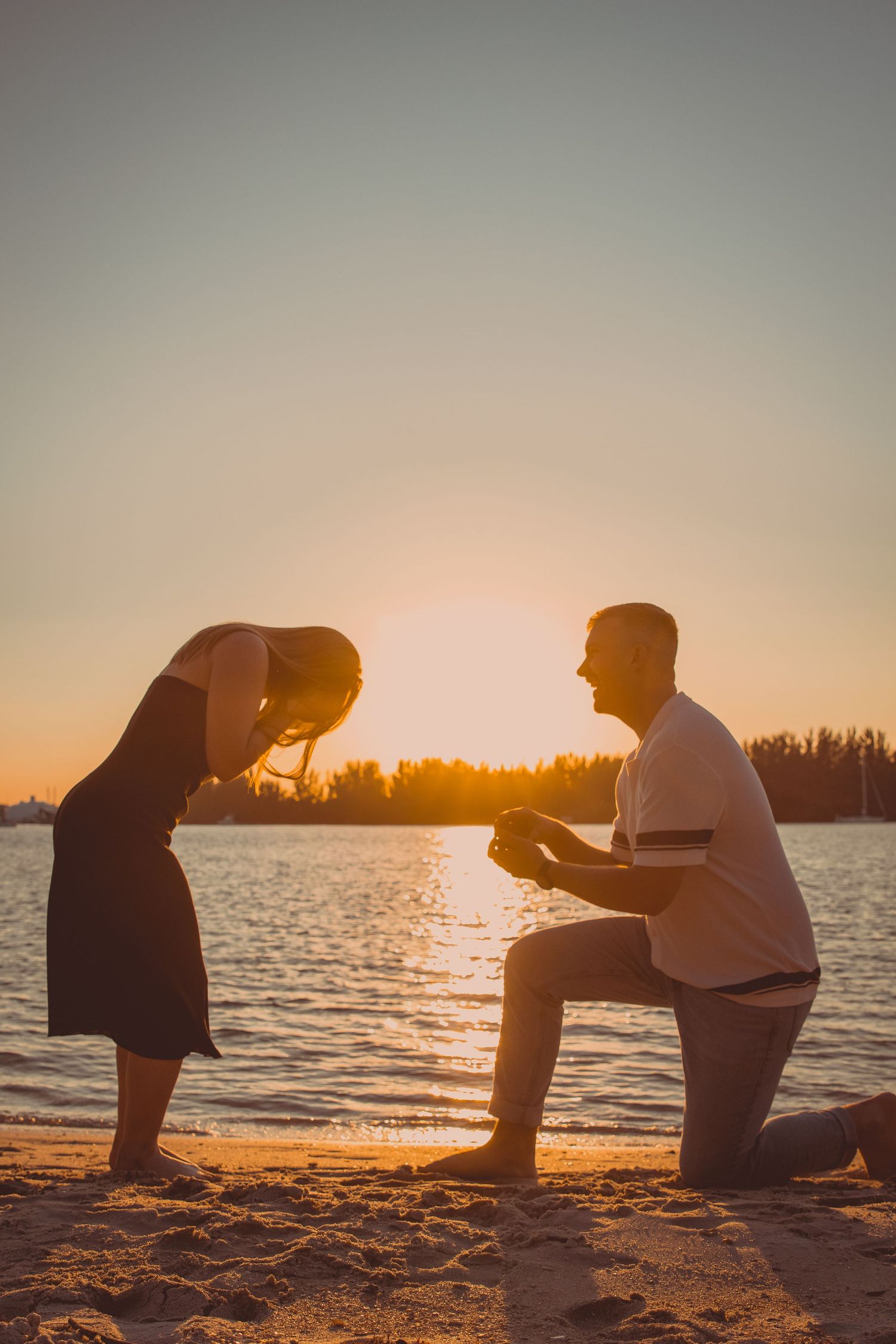 Surprise proposal photography of a man on one knee at sunset as his girlfriend begins to buckle in astonishment.