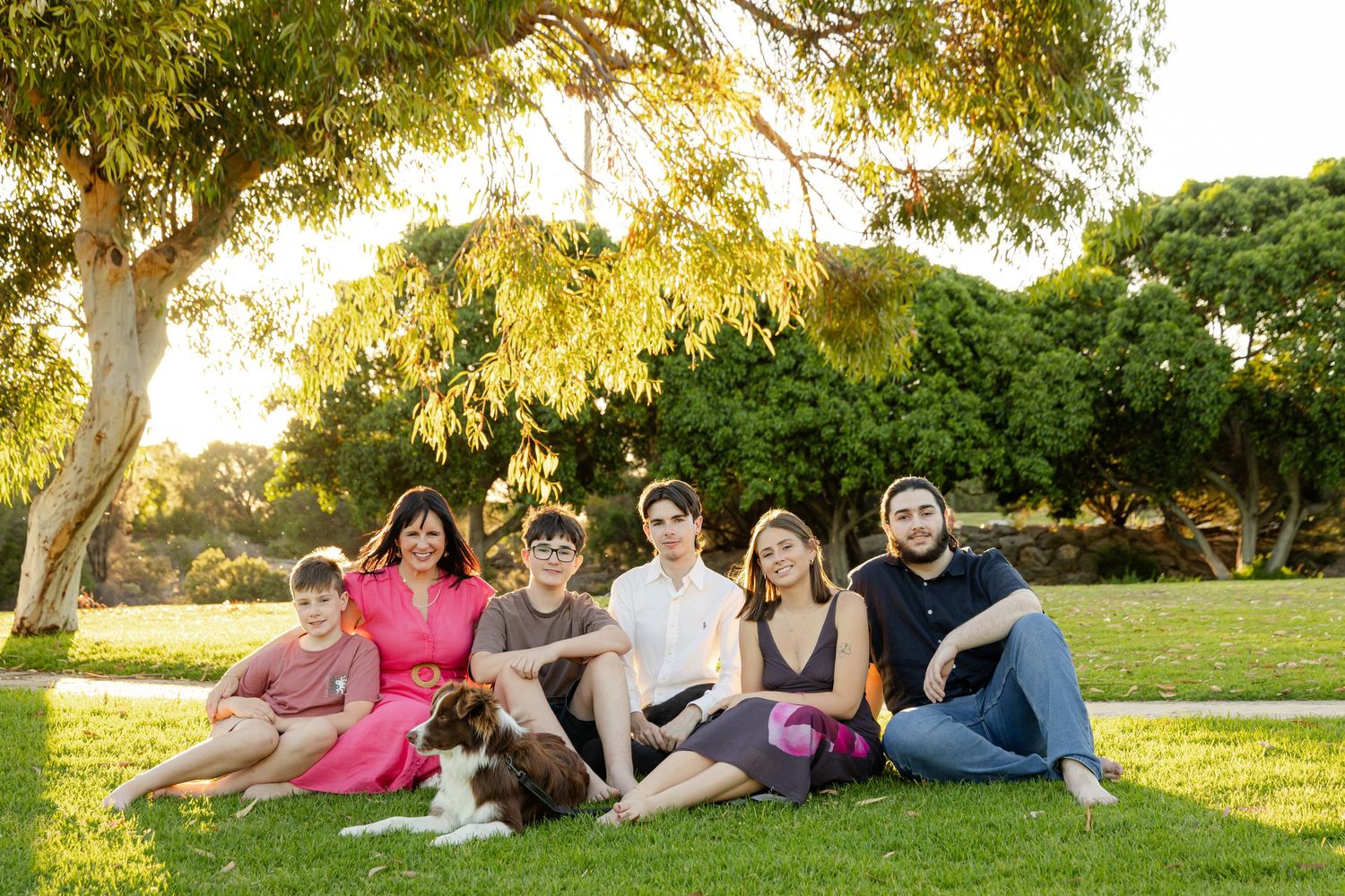 Family of six poses with a dog on green grass in a sunny park with lush trees in the background.
